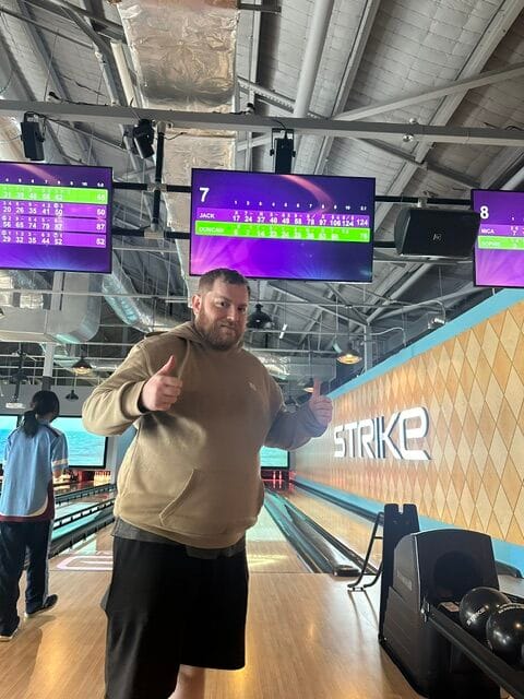 Person giving thumbs up at a bowling alley, relishing a unique day out near lanes with electronic scoreboards overhead.