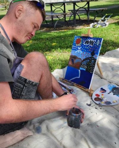 1000104342 Person sitting on a blanket in a park painting on a canvas with a brush, surrounded by painting supplies.