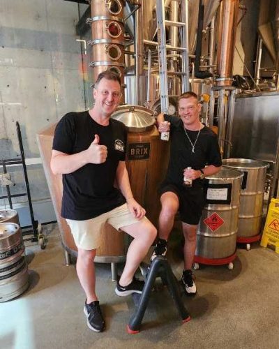 1000111316 Two people posing in front of brewing equipment inside a brewery, both giving thumbs up. One is seated on a stool with a glass in hand; the other is standing beside, smiling.